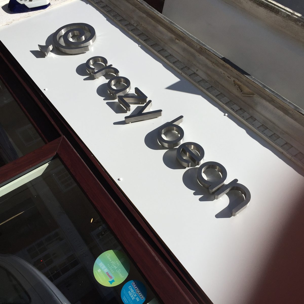 Brushed metal-effect '@cozybean' letters on a white shopfront fascia, steep angle